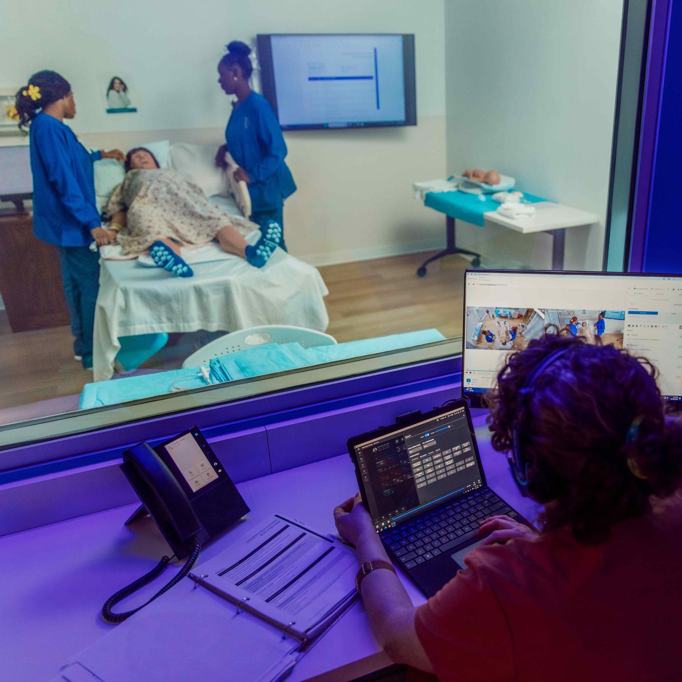 Two students working in simulation lab with instructor in viewing station with technology.