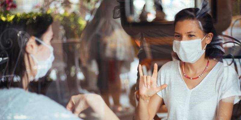 Two women wearing masks, one of them saying "I love you" in American Sign Language