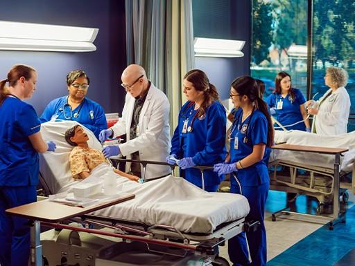 Simulation exam room with mannequin patient, doctor and nursing students surrounding.