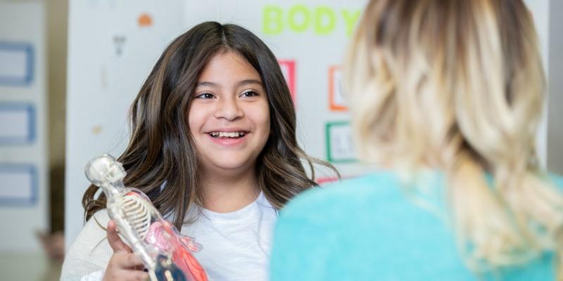 Child holding anatomy skeleton in front of adult in classroom