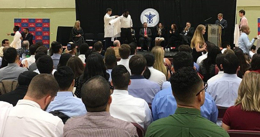 students sitting at ceremony while student on stage receives a white coat